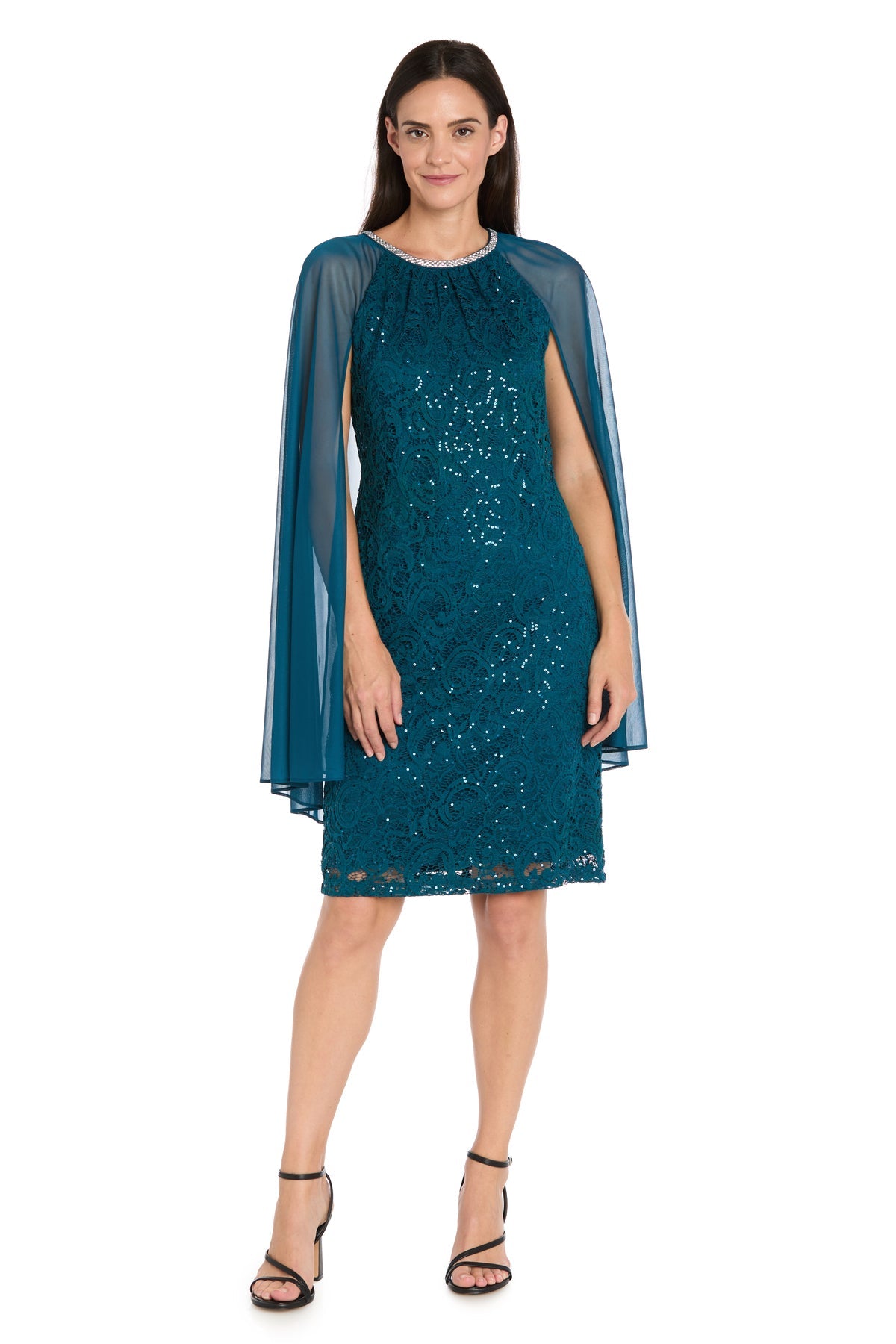 A woman wearing the R&M Richards 3864 Knee-Length Cape Party Dress in teal with sequin lace and a sheer cape overlay, paired with black high-heeled sandals, stands against a white background.